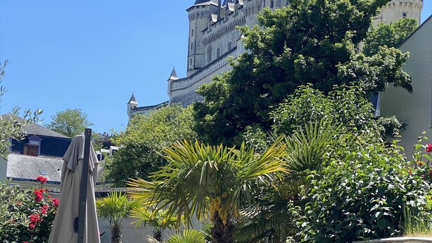 Un patio con una mesa y sillas, rodeado de abundante vegetación y un edificio histórico con torres al fondo.