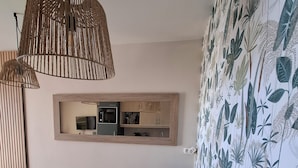 Een eetgedeelte met een tafel, stoelen, een hanglamp, een aan de muur bevestigd spiegel en een keuken op de achtergrond.