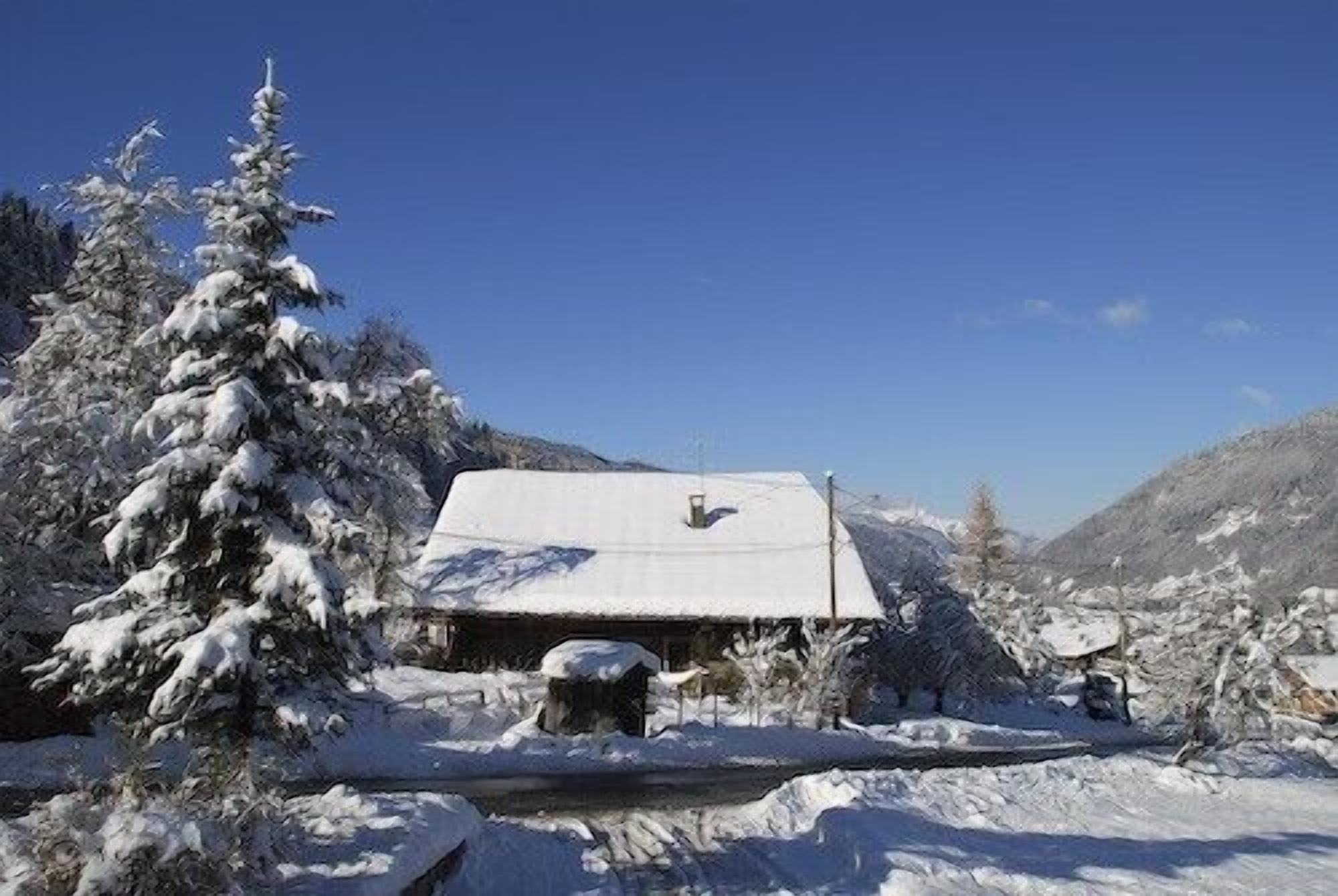Une cabane enneigée, avec un toit en pente, entourée d’arbres chargés de neige et de montagnes en arrière-plan.