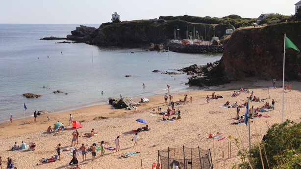 A beach with people sunbathing and swimming, a lighthouse in the background, and a rocky coastline.