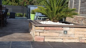 A stone planter with a palm plant, a wooden deck, and a patio area with a table and chairs.