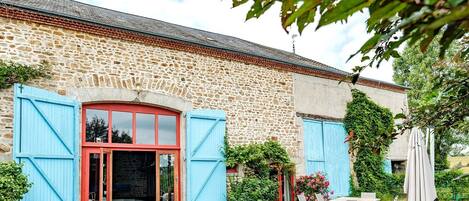 A stone building with a red door and blue shutters, outdoor seating, and a gravel courtyard.