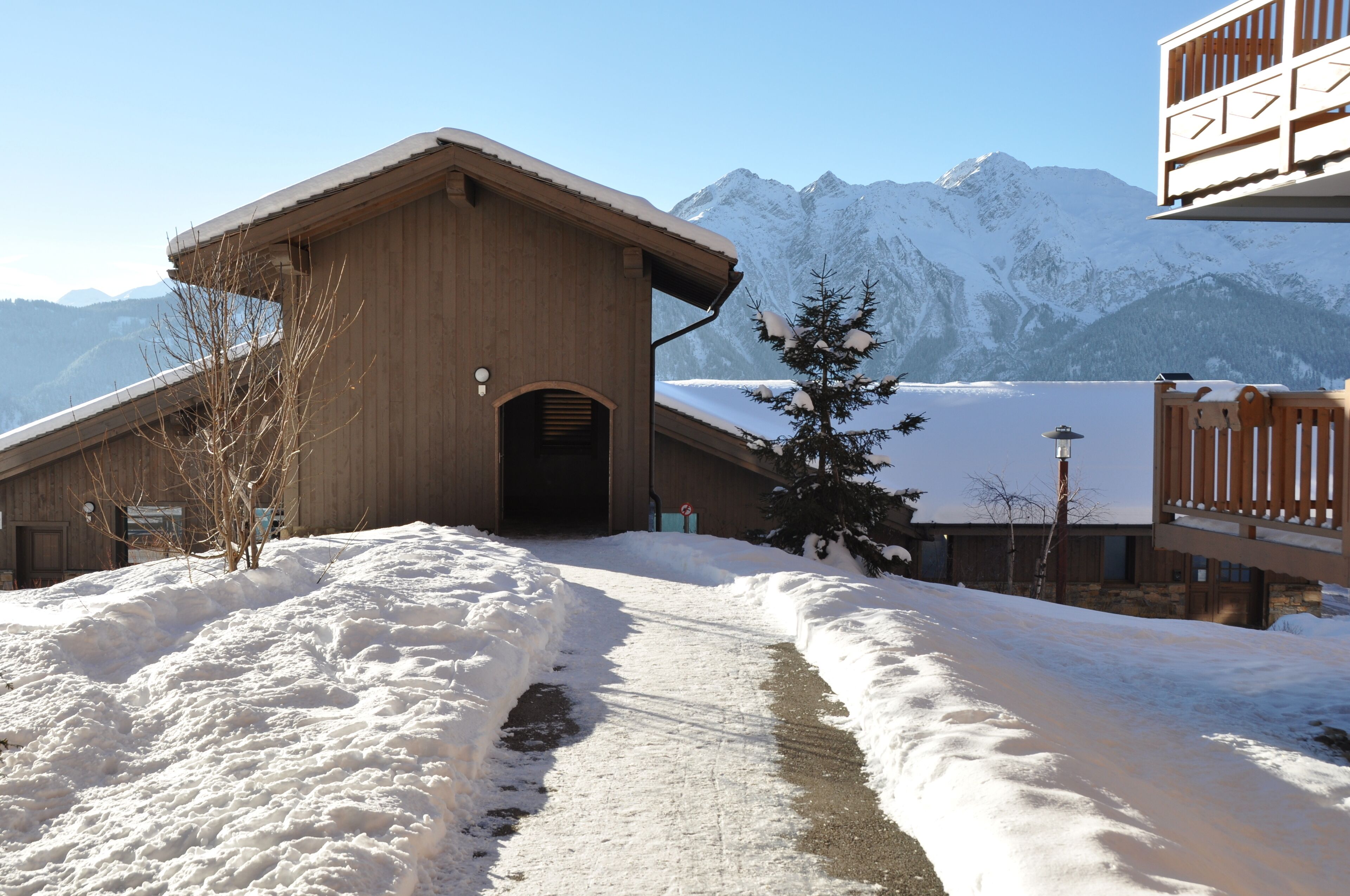 Un edificio de madera con techo inclinado, un sendero cubierto de nieve que lleva a él, y una montaña al fondo.