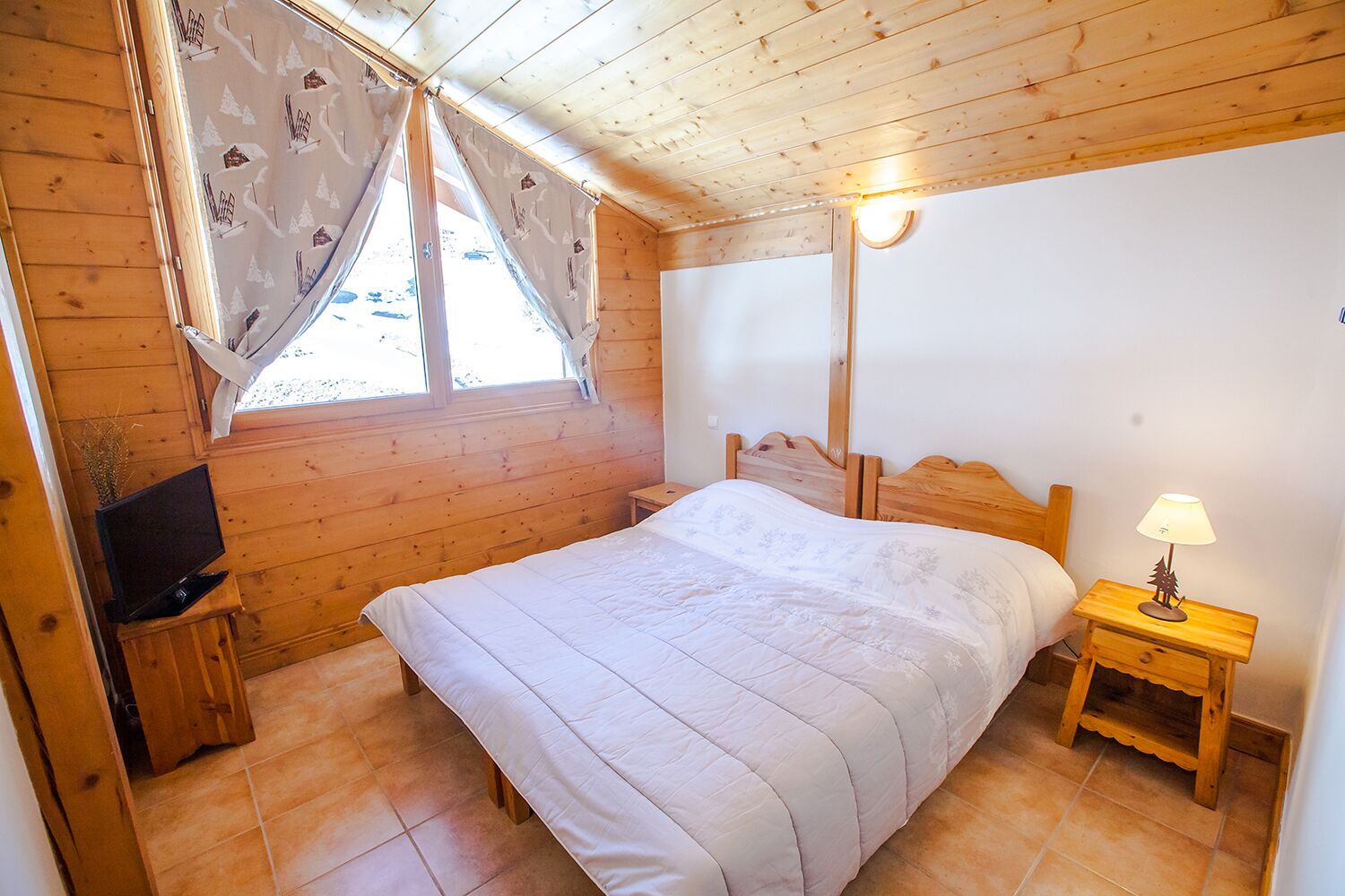 Ein Zimmer mit Holzwänden, einem Bett, einem Fernseher, einer Lampe und einem mit Vorhängen versehenen Fenster.