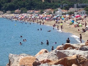 Una playa concurrida, gente nadando y tomando el sol, rodeada de edificios y rocas.