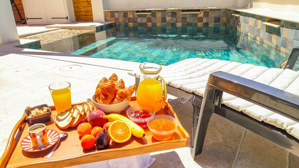 A tray with breakfast items including croissants, fruits, juice, and a cup of coffee by a poolside lounge chair.