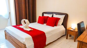 Een net opgemaakt bed met een rood-witte spreidek en twee rode kussens, plus een handdoek in de vorm van een witte zwaan. Er staat een houten nachtkastje met een lamp naast het bed.