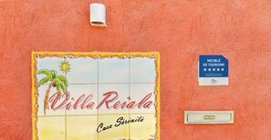 Naamplaatje voor 'Villa Reial Casa Serenità' met een palmpentree- en zonne-illustratie, een blauw bord met sterren en tekst, en een lichtschakelaar aan de muur.
