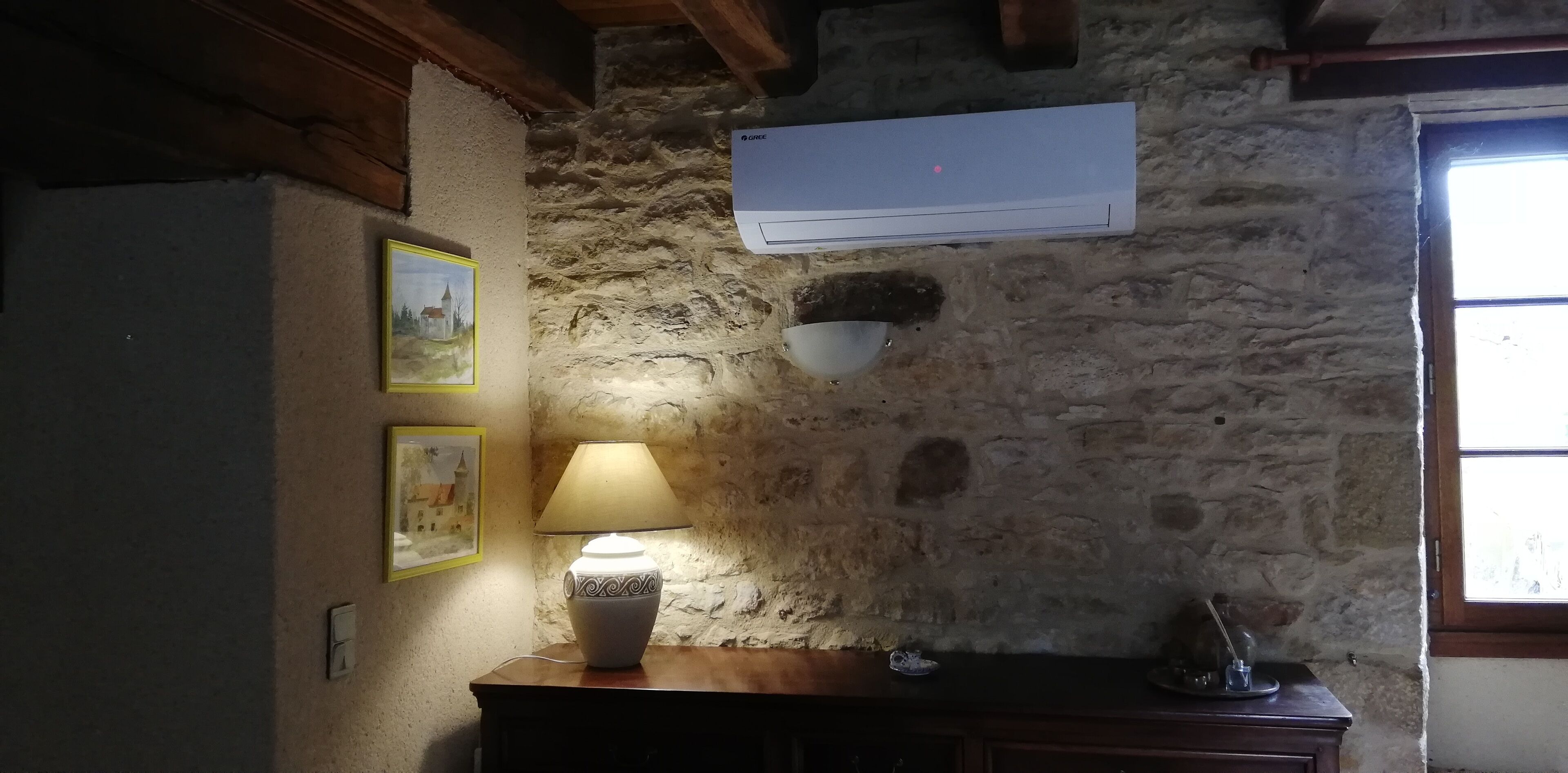 Une pièce rustique avec un mur en pierre, un climatiseur fixé au mur, une table avec une lampe et une fenêtre.