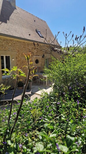 Een stenen huis met een pannendak, een terras met tuinmeubilair en een tuin met verschillende planten en bloemen.
