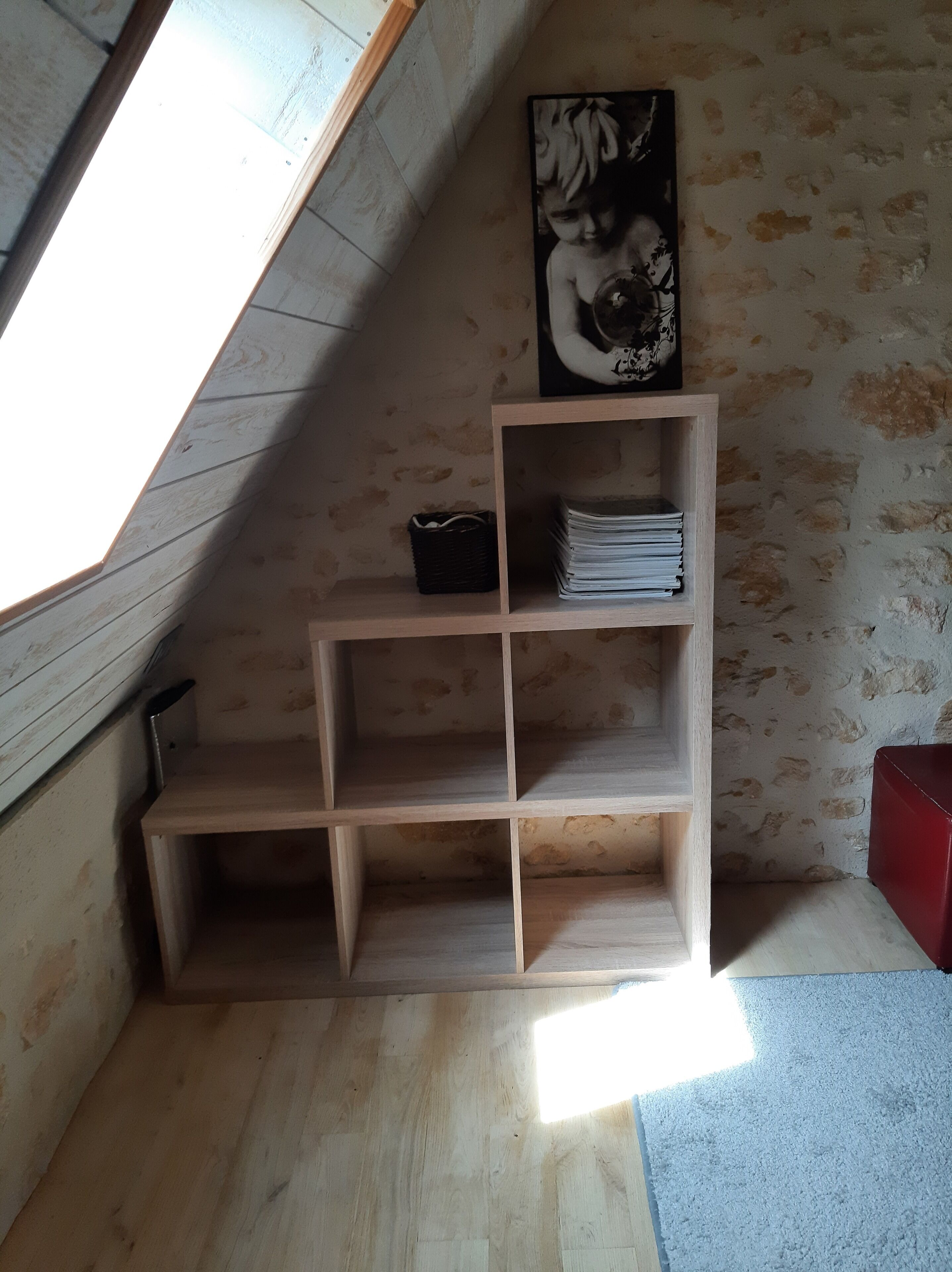 Una estantería de madera con un cuadro enmarcado encima, ubicada en una habitación con techo inclinado y pared con textura.