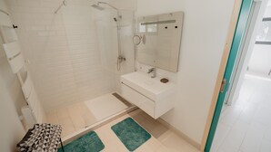 Une salle de bain moderne avec une douche à parois de verre, des carreaux blancs, un lavabo avec un miroir et deux tapis verts sur le sol.