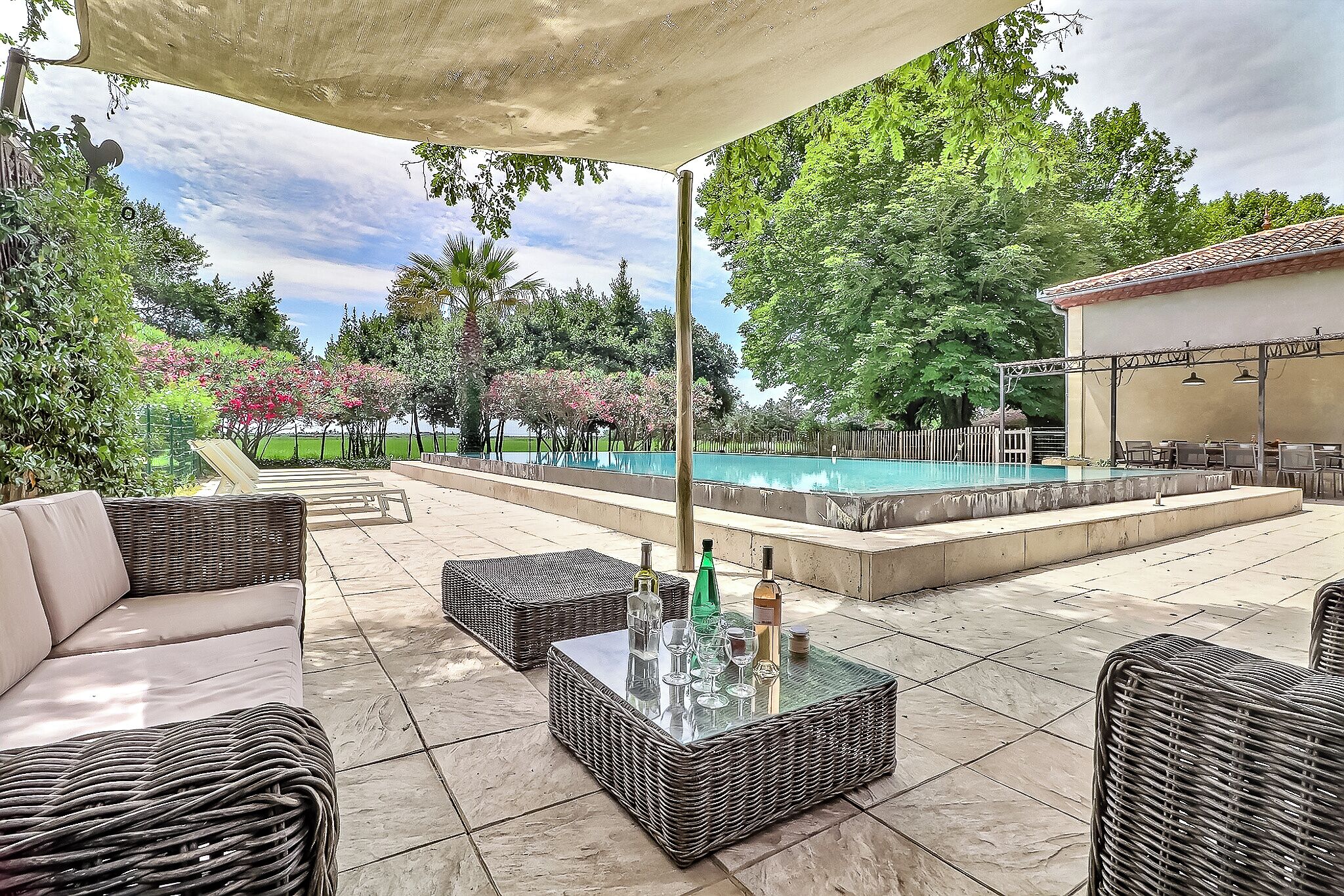 Un espace extérieur aménagé en terrasse, avec des meubles en osier, une table en verre sur laquelle sont posées des bouteilles, et une piscine en arrière-plan.