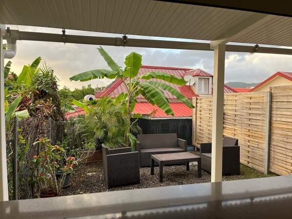 A covered outdoor patio with a seating area, potted plants, and a view of a house with a red roof. - Pleasant apartment. on the ground floor 5 mins walk from the beach, Sainte Anne (SAINTE ANNE)