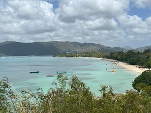 A coastal view with clear turquoise waters, boats, and a sandy beach with a backdrop of hills and a partly cloudy sky.