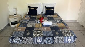 A neatly made bed with a patterned bedspread, two pillows, a nightstand with a lamp, and a painting of a beach scene above the bed. - Pleasant apartment. on the ground floor 5 mins walk from the beach, Sainte Anne (SAINTE ANNE)