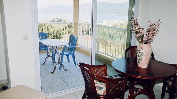 A balcony with a table and chairs, a vase with flowers, and a view of a body of water and hills. - BEAUTIFUL APARTMENT FOR 4 PEOPLE WELL (Propriano)