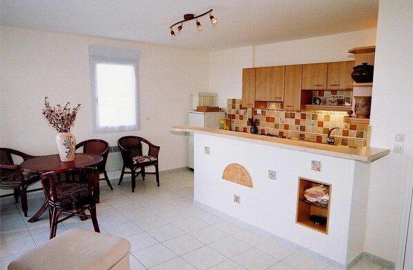 A compact kitchen with wooden cabinets, a tiled backsplash, and a small island. There is a dining area with a table and chairs, and a vase with flowers on the table.