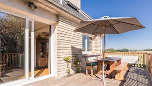A wooden deck with a table and chairs, a large umbrella, and a glass door leading to an interior space. - Beautiful house with sea view and fireplace (Ver-sur-Mer)