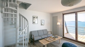 Un salon moderne avec un escalier en colimaçon, un canapé gris, une table basse en bois et une vue sur la mer grâce à une grande porte-fenêtre coulissante.