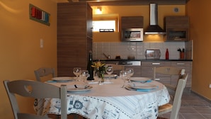 A dining area with a table set for four, a kitchen in the background, and a window providing natural light. - cottage swallows (Riec-sur-Bélon)