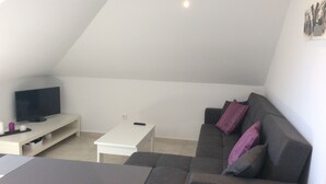 Un salon moderne avec un téléviseur à écran plat posé sur un meuble blanc, un canapé gris agrémenté de coussins violets et une table basse blanche.