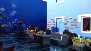 Un espace intérieur avec un mur bleu et une fresque représentant la vie marine. On y trouve plusieurs tables et chaises, dont certaines sont ornées de plantes en pot.