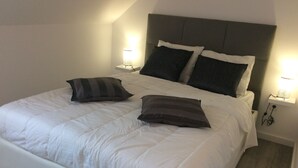 Un lit bien fait, avec une literie blanche et deux oreillers, une tête de lit grise et des lampes de chevet de chaque côté.