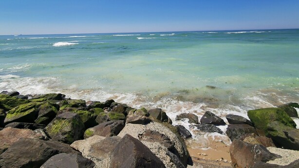 A beach with clear turquoise water, waves, and large rocks in the foreground.