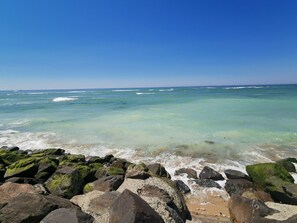 A beach with clear turquoise water, waves, and large rocks in the foreground.