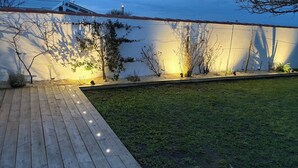 A wooden deck with string lights, a grassy yard, and a white wall with climbing plants.