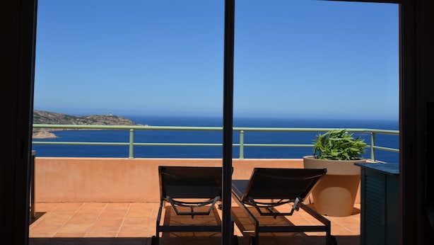 A balcony with two chairs and a potted plant, overlooking a coastal view with the sea and distant hills.