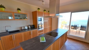 A modern kitchen with wooden cabinets, a large island, and a view of the ocean through the sliding glass doors.