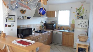 Une cuisine avec des meubles en bois, un four à micro-ondes, une cuisinière, un lave-vaisselle et un réfrigérateur. On y trouve également des filets suspendus et une chaise en bois.