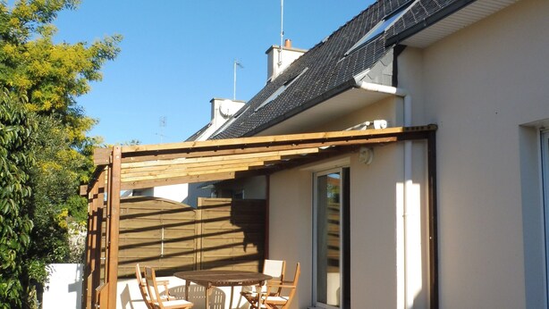 Une terrasse avec une table et des chaises, une pergola en bois et une maison avec un toit en tuiles.