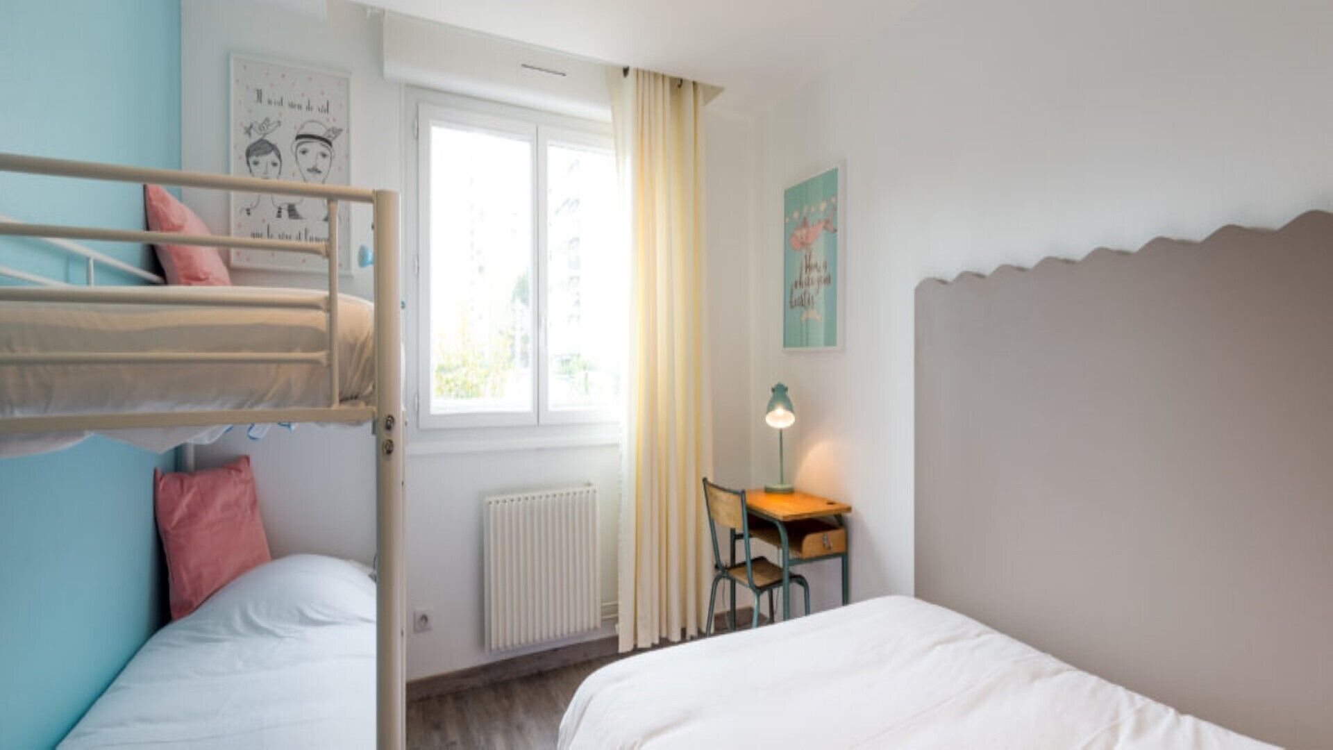 A room with bunk beds, a desk with a lamp, a radiator, and a window with curtains.
