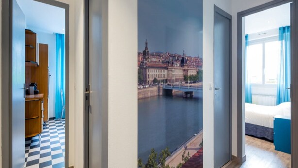Ein Hotelzimmer mit Schachbrettboden, einem Bett, einem Kleiderschrank und Blick auf eine Stadtlandschaft mit einer Brücke über einem Fluss.