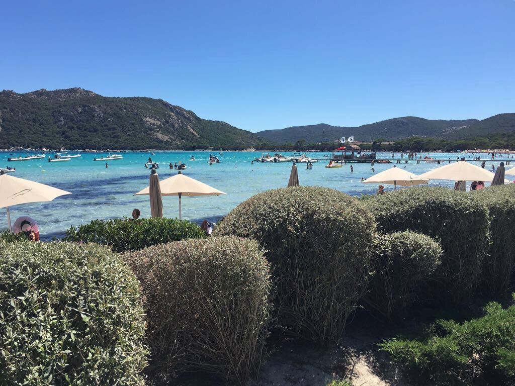 En strand med kristallklart blått vatten, båtar och parasoller.