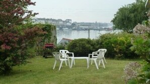 A grassy outdoor area with two white plastic chairs and a small table, overlooking a body of water with boats and buildings in the background. - South Brittany House Sea View in Port-louis 8 -10 people (Port-Louis)