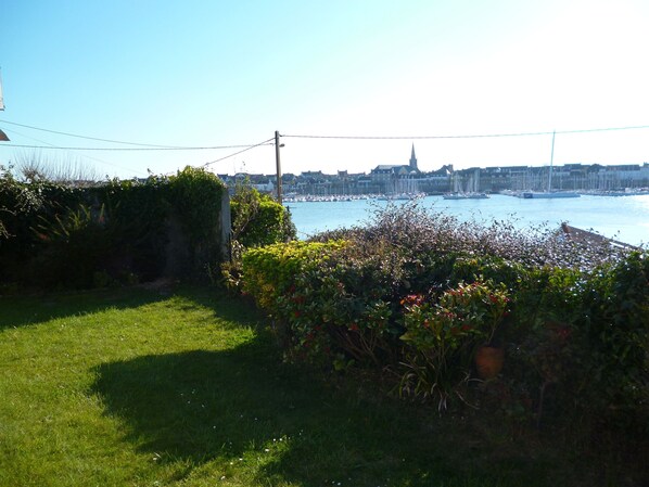 A coastal view with a grassy area, dense shrubbery, and a distant marina with boats.