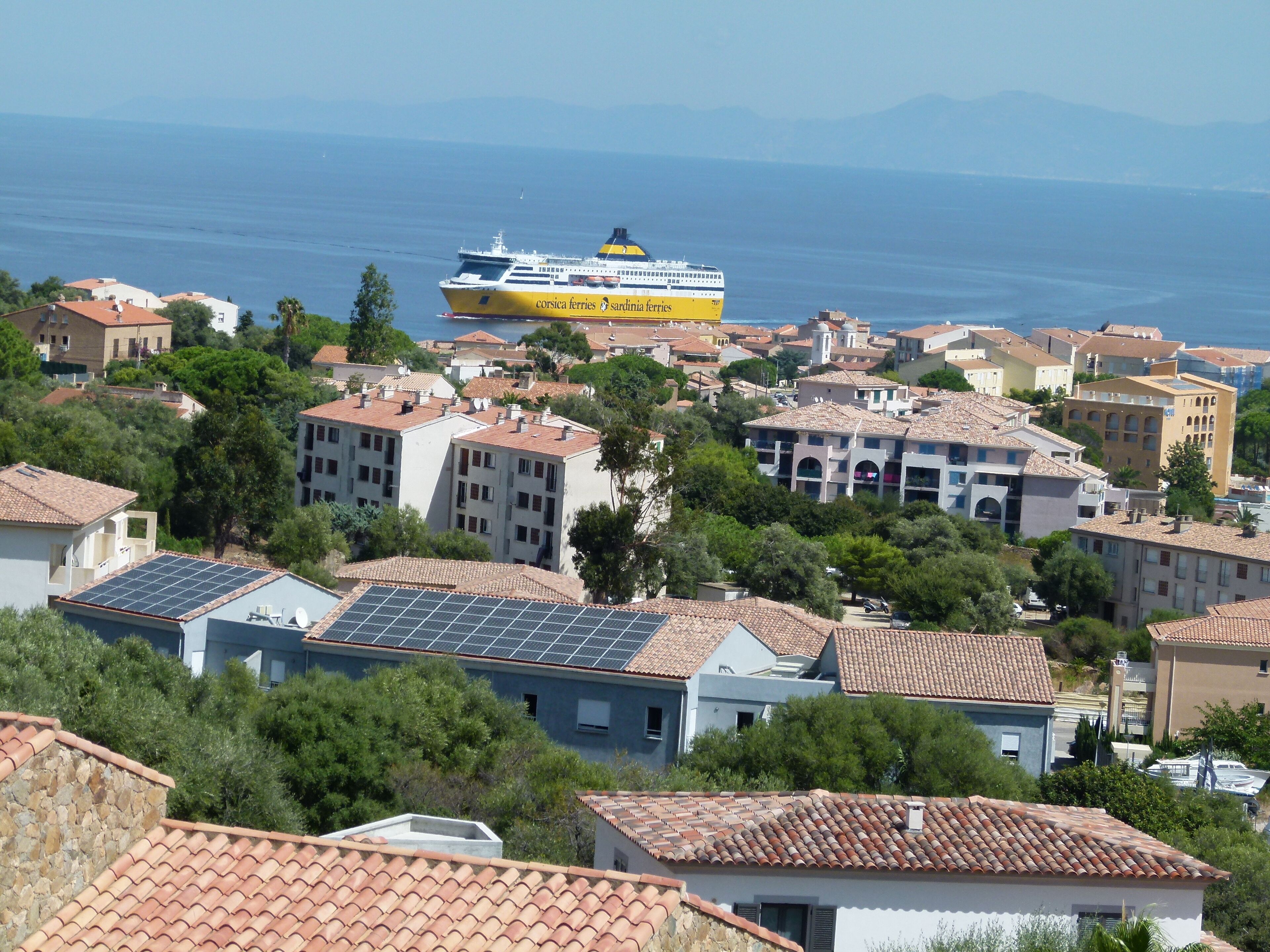 Un pueblo costero con arquitectura mediterránea, que incluye edificios con tejados de tejas rojas y un ferry al fondo.