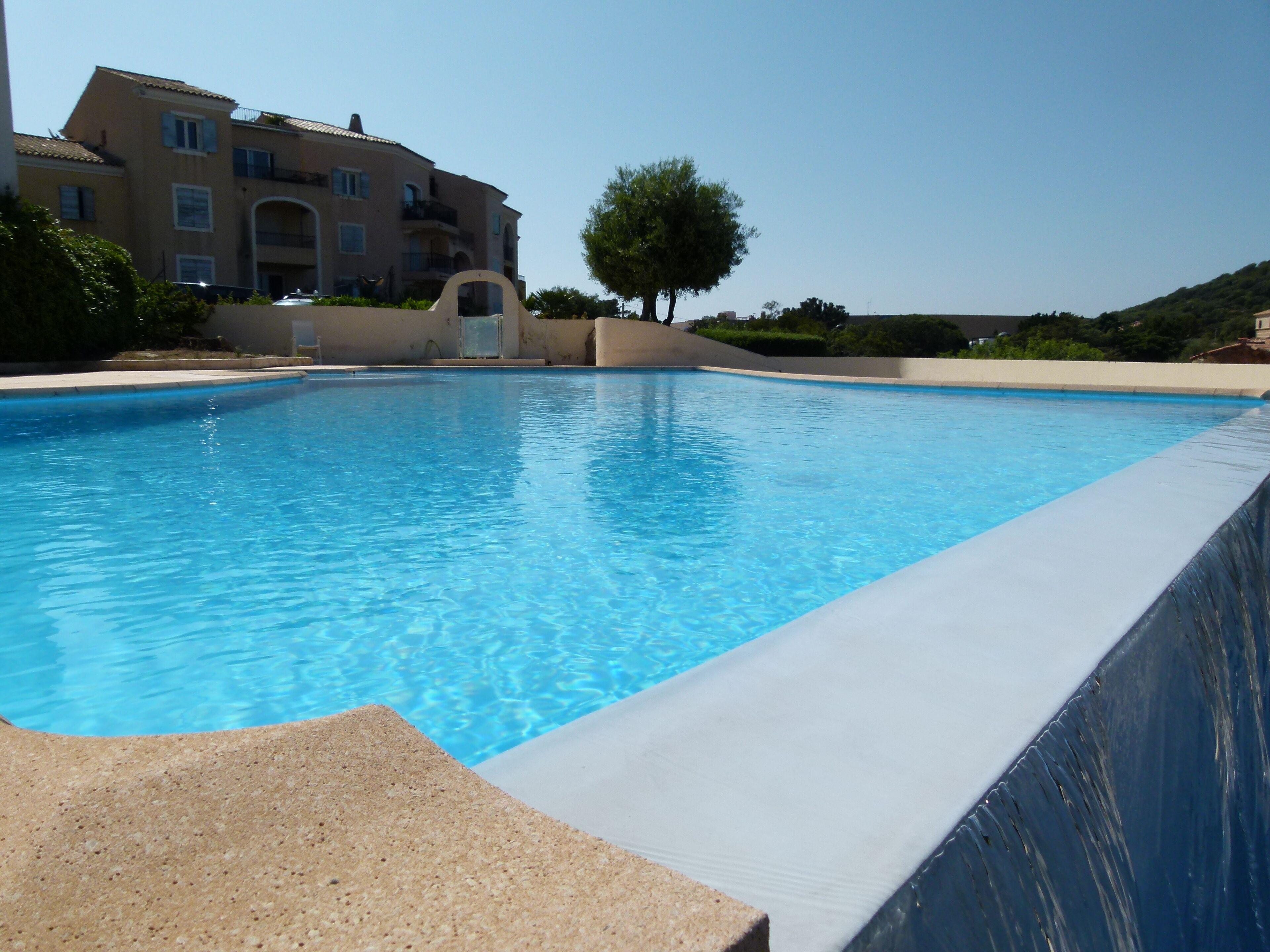 A swimming pool with a clear blue water surface, surrounded by a low concrete wall. In the background, there is a multi-story building with balconies and a tree.