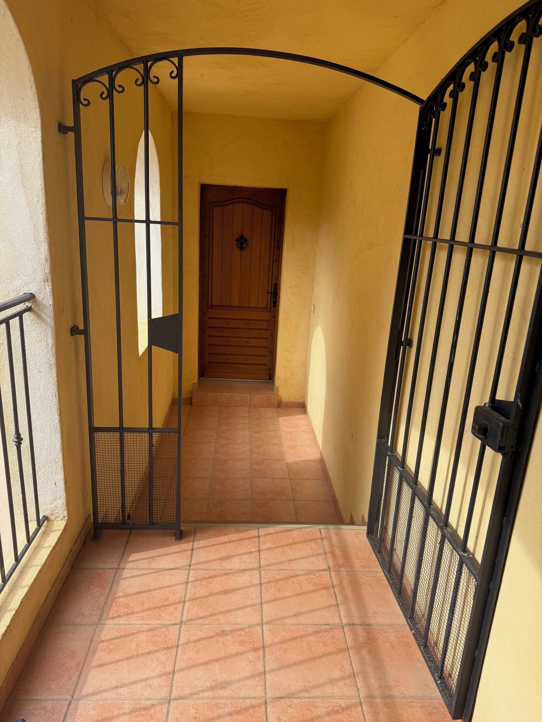 Un couloir se terminant par une porte en bois, de part et d’autre de laquelle se dressent des grilles en métal noir.