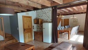 A cozy living room with a wooden sofa, a small wooden table, a stone wall, a white refrigerator, and a dining area with a long wooden table and chairs. - Gîte 25 p, swimming pool, whirlpool, fitness, sauna ⚠️Vérifiez spams⚠️ (Charensat)