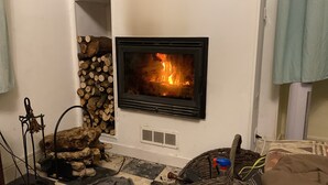 A fireplace with a visible fire, a stack of firewood, and a radiator. - VILLA FOR RENT (near the beach with swimming pool, garden barbecue). (Menton)
