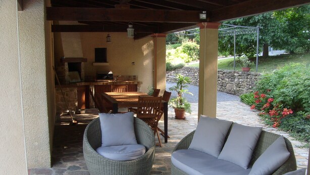 A covered patio area with wicker furniture, a stone floor, and a view of a garden with a stone wall and potted plants. - Quiet, restful house with private pool in a tourist area (Bout-Du-Pont-De-Larn)