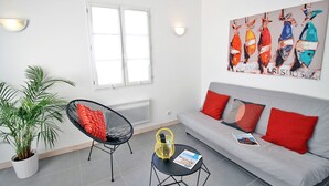 Ein modernes Wohnzimmer mit einem grauen Sofa, roten Kissen, einem schwarzen Sessel, einem kleinen Tisch mit einer Zeitschrift, einer Zimmerpflanze und einem Bild bunter Fische an der Wand.