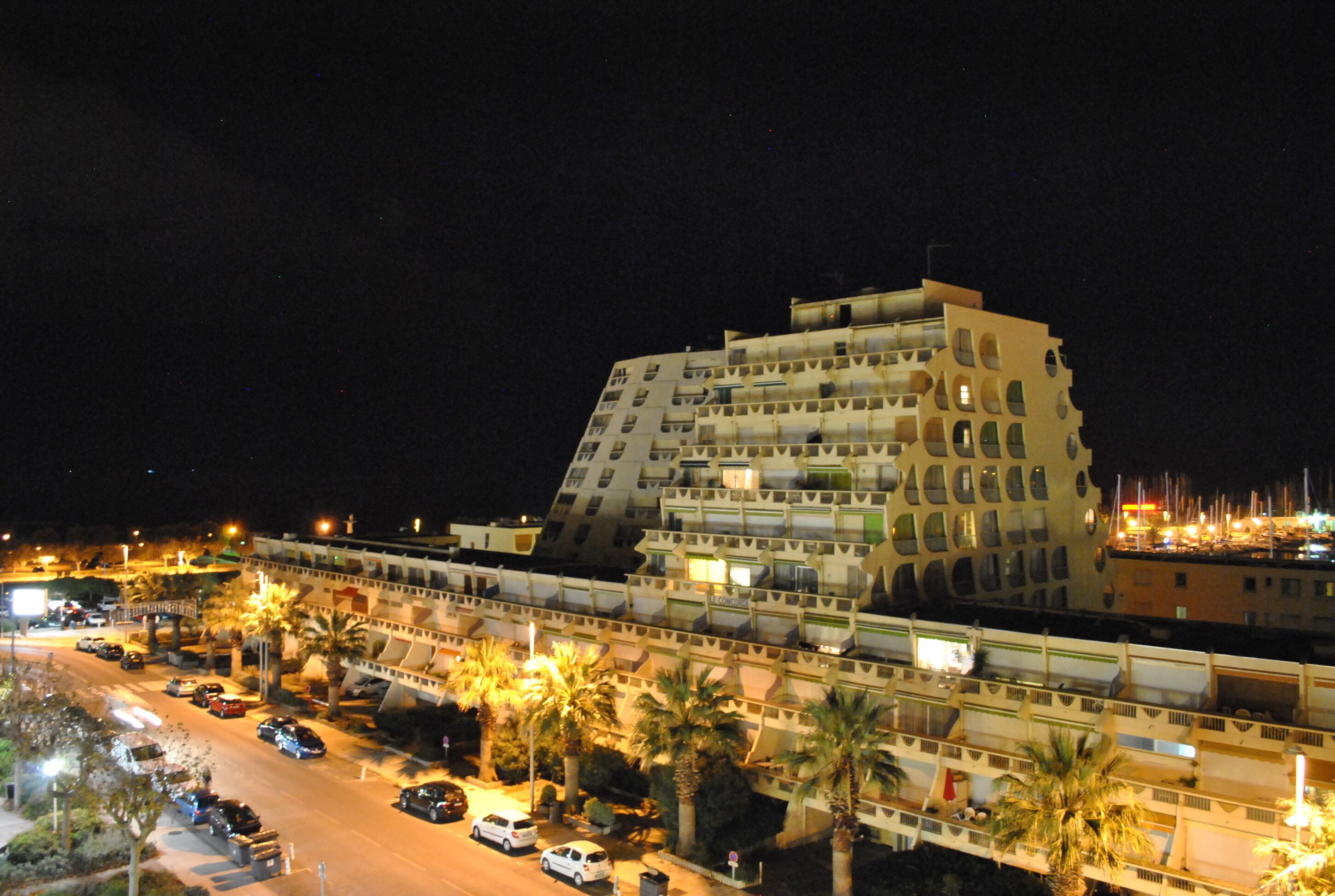 Un hotel de varios pisos con un diseño arquitectónico único, con balcones y un estacionamiento que incluye coches y palmeras.
