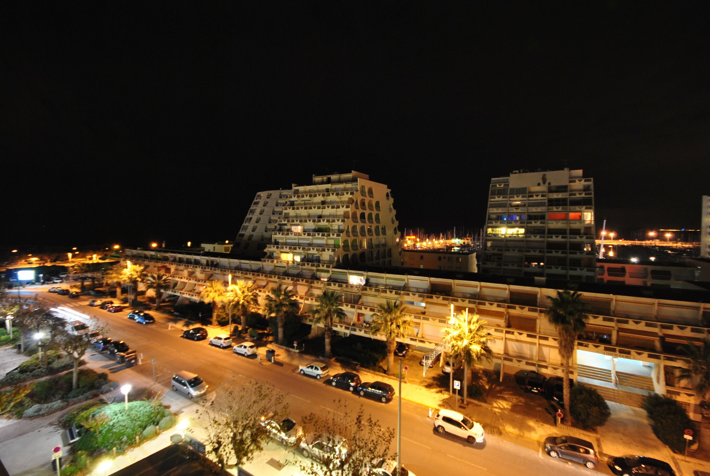 Une vue nocturne d’une ville avec ses bâtiments modernes, une rue bien éclairée et des voitures garées.