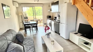 A modern living room with a grey sofa, a white coffee table, and a TV stand. There is a kitchen area with a washing machine and a dining area with a table and chairs. - T2 apartment, 40 m2, fully air-conditioned, 350 m from the beach. Private parking (Saint Cyr sur Mer)